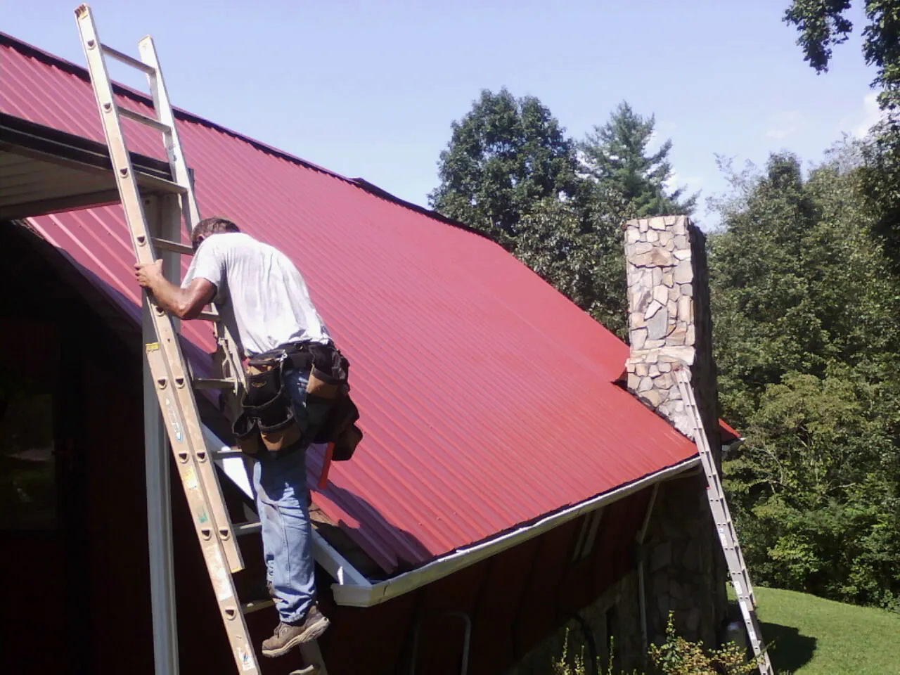Roofer on a ladder beside a red metal roof and stone chimney — Soffit & Fascia Repair by Roofing Brothers in Potomac